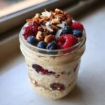 A glass jar filled with creamy overnight oats, topped with fresh raspberries, blueberries, nuts, and coconut flakes.