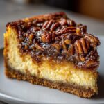 A decadent slice of Pecan Pie Cheesecake on a white plate, showcasing a creamy cheesecake layer topped with glossy pecan pie filling.