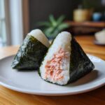 Two salmon Onigiri rice balls wrapped in nori seaweed on a white plate. One is cut open to show the salmon filling.