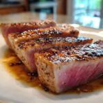 Close-up of four perfectly seared ahi tuna steaks, cooked rare to medium-rare, glistening in a savory sauce.