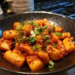 Close-up of Tteokbokki, Korean spicy rice cakes, in a black bowl, garnished with green onions.