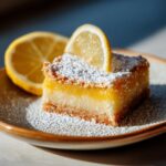 A single vegan lemon bar slice on a plate, topped with powdered sugar and a lemon slice.
