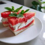 Two watermelon sandwich pieces with cream cheese filling and mint garnish on a white plate.