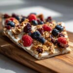 A close-up of homemade yogurt bark topped with fresh blueberries, raspberries, granola, and dried cranberries.