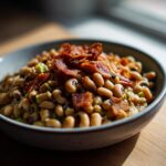 A bowl of black eyed peas recipe, garnished with crispy bacon pieces, sits on a wooden surface.