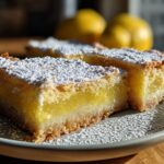 Close-up of lemon bars 9x13 pan, sliced and dusted with powdered sugar on a plate with lemons in the background.