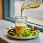 Lemon vinaigrette dressing being poured into a jar, surrounded by fresh salad greens.