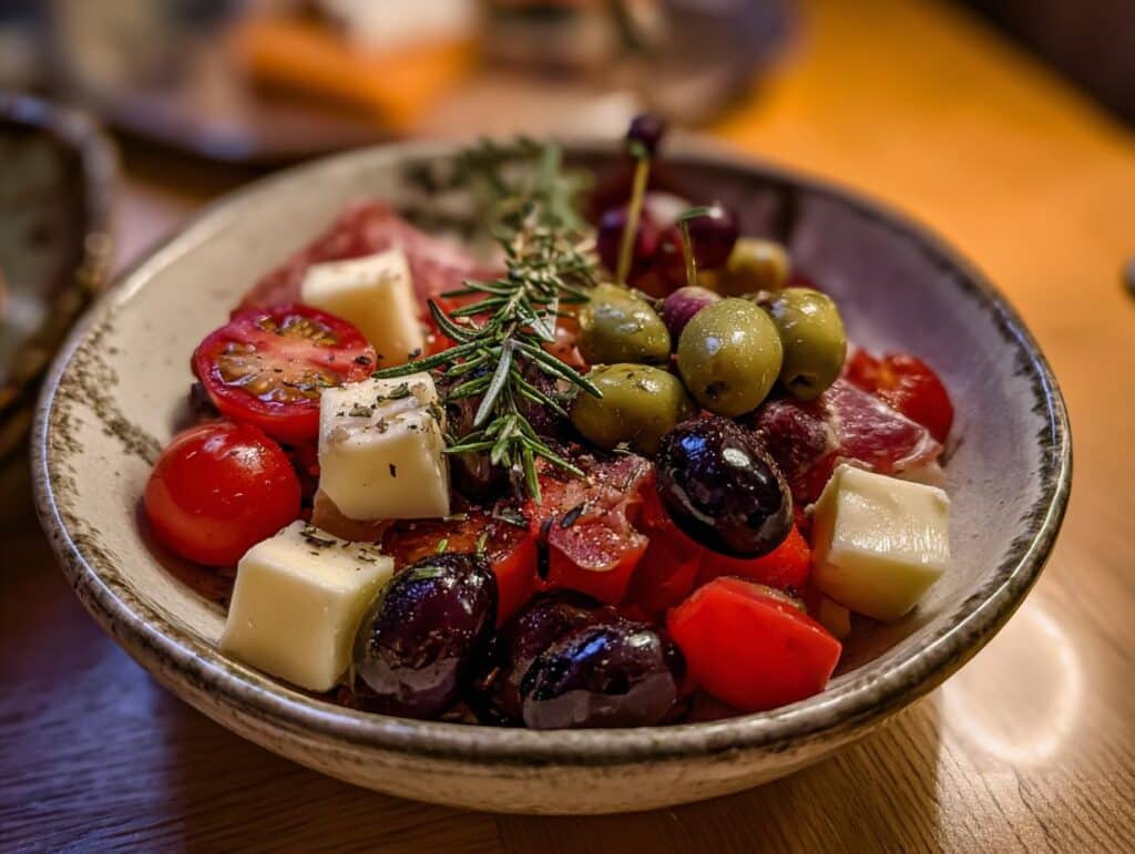 A bowl of Antipasto Salad with tomatoes, olives, cheese, and salami, garnished with fresh rosemary.