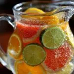 Close-up of a glass pitcher filled with clear water, sliced lemons, limes, grapefruits, and oranges, with visible bubbles. Citrus Infused Water Recipe.