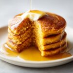 A stack of classic sweet potato pancakes with a pat of butter and maple syrup being poured over them, one pancake cut open.