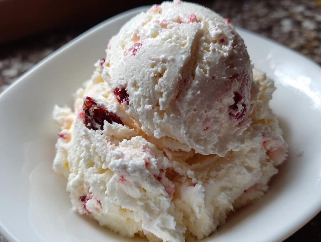 A scoop of creamy Cottage Cheese Ice Cream with red fruit pieces in a white bowl.