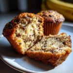 A close-up of a Banana Protein Muffin split in half, revealing a moist interior with abundant chocolate chips on a plate.