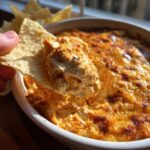 A tortilla chip being dipped into creamy buffalo chicken dip, showing the texture and color of the dip.