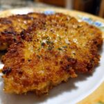 Close-up of a golden-brown, crispy chicken breast recipe on a plate, seasoned with herbs.