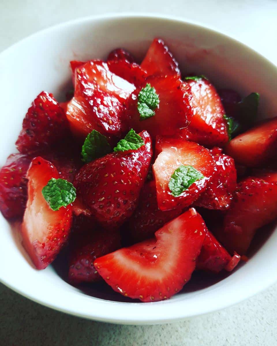 Close-up of a white bowl filled with sliced fresh strawberry recipes, garnished with mint leaves.