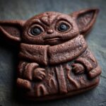 A Grogu-shaped baby in bloom cookies, dark brown, on a dark gray slate background.