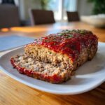 A sliced meatloaf topped with a vibrant tomato glaze and fresh herbs, sitting on a white plate.