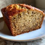 Close-up of a moist banana bread loaf on a white plate, showcasing its texture and golden-brown crust.