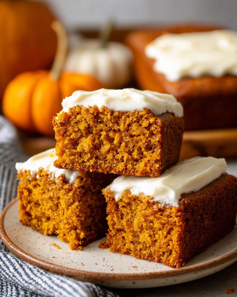 Three stacked pumpkin bars with white cake mix easy, topped with white frosting, on a plate with pumpkins in the background.