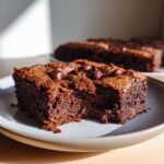 A close-up of a slice of sourdough brownies recipe, topped with chocolate chips, on a plate with a bite taken out.