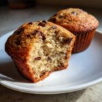 Two bakery style chocolate chip muffins, one with a bite taken out, showing the soft interior.