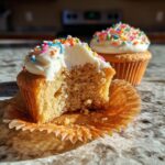 Two easy spring cupcakes, one with a bite taken out, showing the fluffy interior and creamy frosting with colorful sprinkles.