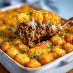 Serving a cheesy tator tot casserole with ground beef and veggies from a white baking dish.