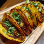 A row of flavorful Birria Tacos on a wooden board, garnished with cilantro and onions.