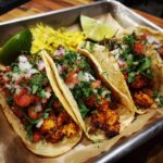 Three Chicken Street Tacos on a tray with lime wedges and toppings like cilantro, onion, and salsa.