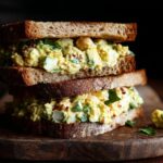 Two chickpea salad sandwiches stacked on a wooden board, showcasing the creamy filling and whole wheat bread.