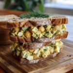 Two chickpea salad sandwiches stacked on a wooden board, garnished with dill. Delicious chickpea salad sandwiches.