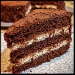 A decadent slice of The Chocolate Cake with layers of chocolate cake and cream filling, topped with cocoa powder.