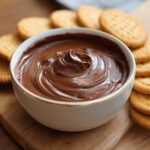 A bowl of Velvety 5-Minute Chocolate Cream Cheese Dip surrounded by crackers on a wooden board.