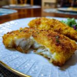 Two pieces of Crispy Baked Parmesan Crusted Tilapia on a plate, one cut open to reveal the flaky fish inside.