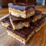 A stack of Easy No-Bake Chocolate Peanut Butter Fudge squares on a wooden board.