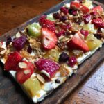 A vibrant The Frozen Yogurt Bark topped with fresh strawberries, pineapple, nuts, and dried fruit on a wooden board.