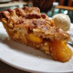 A slice of Grandma's The Peach Cobbler served with a scoop of vanilla ice cream on a white plate.