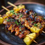 Close-up of Grilled Pineapple Chicken Skewers on a dark plate, garnished with herbs.