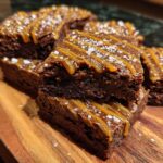 A stack of The Miso Caramel Brownies drizzled with caramel and sprinkled with sea salt on a wooden board.