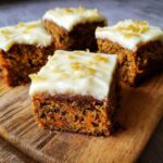 Four moist 350 calorie carrot cake bars with white frosting and candied ginger, sitting on a wooden board.