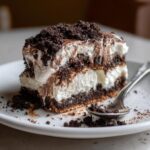 A slice of ultimate no-bake dirt cake with layers of cream and crushed chocolate cookies, served with a spoon.