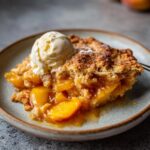 A slice of The Peach Cobbler topped with vanilla ice cream on a plate, golden crust and juicy peaches.