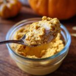 A spoonful of The Pumpkin Fluff is held over a glass bowl filled with the dessert. Pumpkins are in the background.