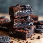 A stack of three Ultimate Fudgy Oreo Brownies, topped with Oreo pieces, on a wooden surface.